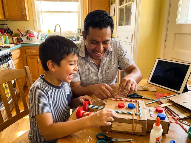A father and son laugh as they build a cardboard robot at the kitchen table